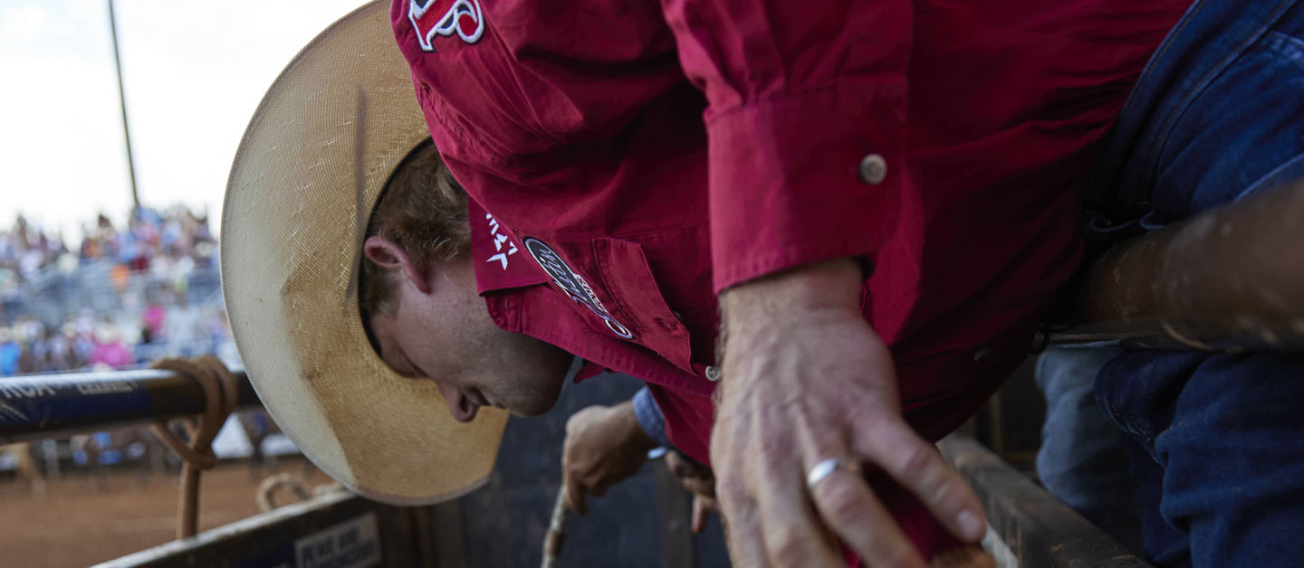 Cowboy wearing a red shirt standing on the chutes putting his bull rope on the bull.